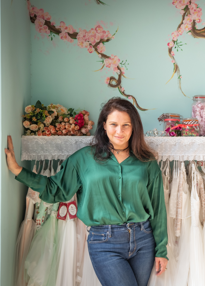 תמונה של נטעלה עומדת בחנות בחולצה ירוקה על רקע קוטלב שמלות בת מצווה וציור קישר של פרחים